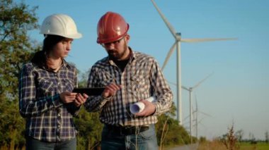 İki mühendis bilgi tablet üzerinde tartışmak. Bir adam bir büyük kağıt rulo tutar. Rüzgar jeneratörleri üzerinde arka plan vardır.