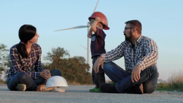 Een gelukkige familie zit op een weg. Een kind speelt met de constructor helmen, kussen moeder en high-fives vader — Stockvideo