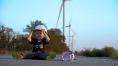 Bir çocuk bir yolda bir arka plan üzerinde rüzgar jeneratörleri ile oturan inşaat kask ile oynuyor. Ağır çekim