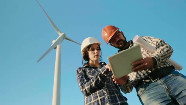 Two engineers in a field are discussing while looking at the tablet. The wind generator is on the background. Slow motion — Stock Video