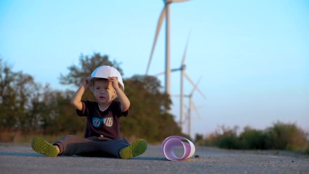 Een kind speelt met de bouw helm, zittend op een weg met windgenerators op een achtergrond. Slow motion — Stockvideo