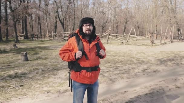 Porträt eines Touristen mit großem Touristenrucksack, mit Brille, Wintermütze und knallroter Jacke am Waldrand — Stockvideo