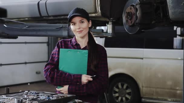 Mecânico de carro menina no fundo do carro em um boné preto e uma camisa xadrez e com um tablet para documentos — Vídeo de Stock