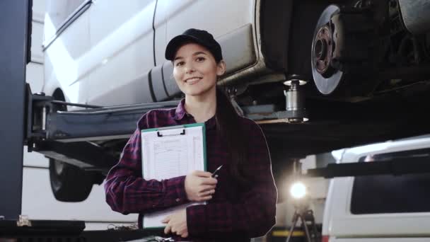 Πορτρέτο του ένα κορίτσι, μηχανικός αυτοκινήτων σε ένα καρό πουκάμισο και ένα μαύρο καπάκι, μπροστά από ένα αυτοκίνητο σε ένα πρατήριο με έγγραφα σε τα χέρια, ευγενικά χαμόγελα και φαίνεται στην κάμερα — Αρχείο Βίντεο