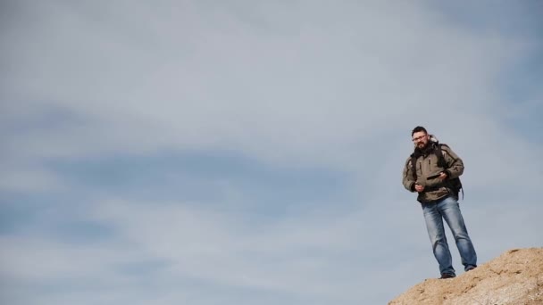 En ung man, en resenär, står på en hög lutning mot bakgrund av en charmig himmel och använder sin mobiltelefon för läggning på terräng. — Stockvideo