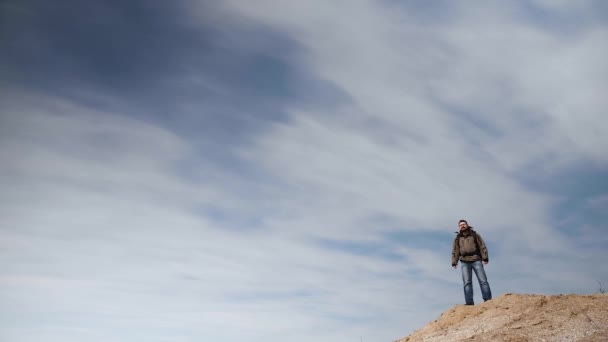 Un joven en equipo turístico se encuentra en una alta pendiente arenosa sobre el telón de fondo de un cielo azul hechizante, que ocupa más espacio en el marco . — Vídeos de Stock