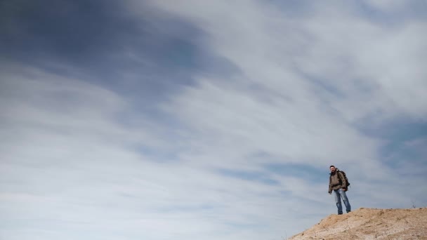 Un giovane uomo in marcia turistica si erge su un alto pendio sabbioso sullo sfondo di un cielo azzurro ammaliante, che occupa più spazio nella cornice . — Video Stock