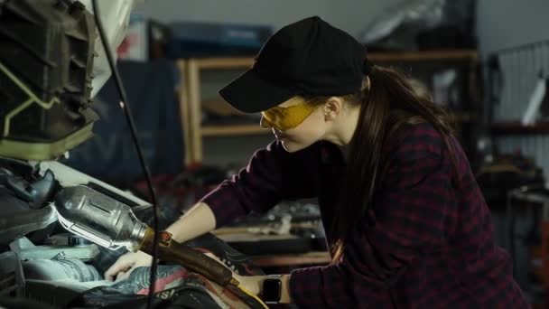 Красива дівчина-механік, брюнетка, в плетеній сорочці і кепці, в захисних окулярах, що ремонтують автомобільний двигун. Концепція рівності чоловіків і жінок — стокове відео