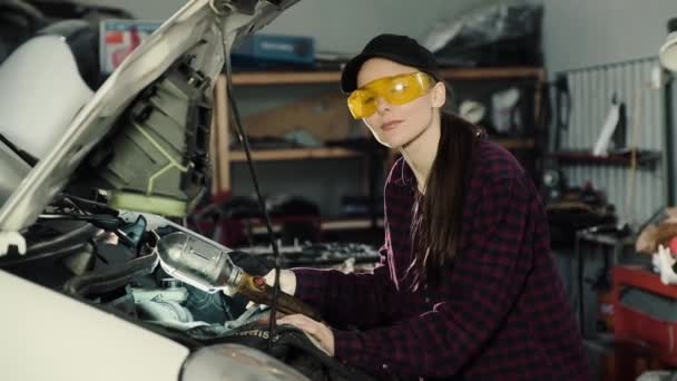 Hermosa morena, mecánica automotriz, con gorra y camisa a cuadros, arregla el motor del coche y mira a la cámara con una sonrisa — Vídeos de Stock