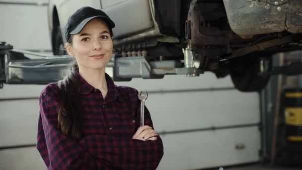 Portret pięknej dziewczyny, mechanika, z kluczem w dłoni na tle samochód na podnośniku — Wideo stockowe