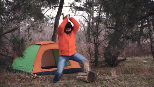 En resenär i ljusa kavaj och vintermössa klipper ved för att göra en brand på bakgrunden av en turist tält för övernattning i skogen. Slow motion — Stockvideo