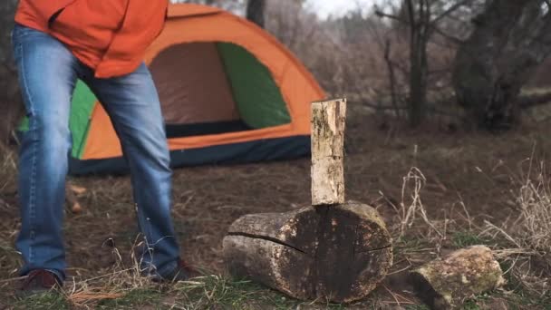 鮮やかなジャケットと冬帽子の旅行者は、森で一晩観光テントの背景に、火災を作るための薪をカットします。スローモーション — ストック動画