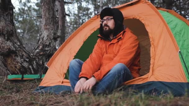 Молодой человек в яркой туристической куртке, в зимней шляпе и очках, сидит у входа в недавно установленную туристическую палатку и наслаждается единством с природой и свежим лесным воздухом . — стоковое видео