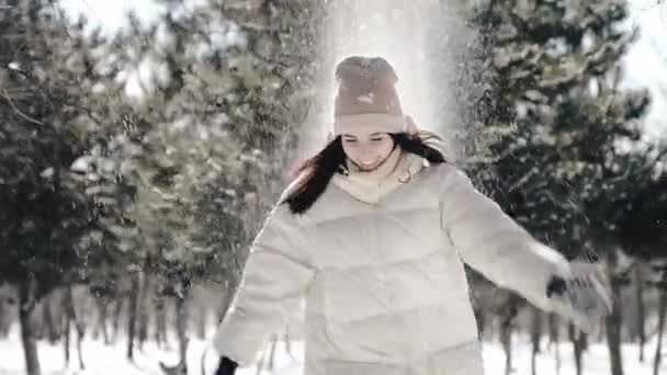 Красива дівчина в зимовому парку весело грає зі снігом, вона кидає обладунки снігу над головою, який пізніше розсипається і перетворюється на багато маленьких сніжинок, що дає багато радості — стокове відео