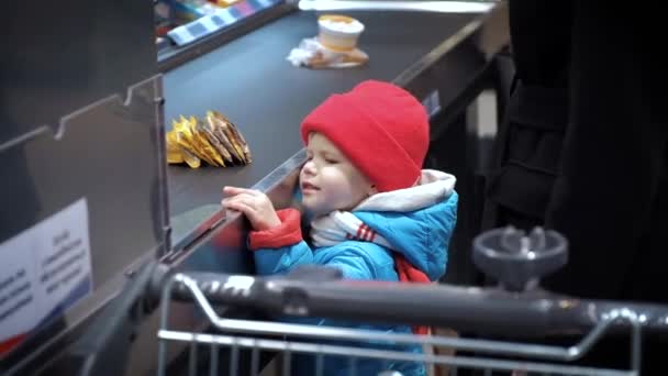 Un niño se para cerca de la caja del supermercado y mira las mercancías — Vídeos de Stock