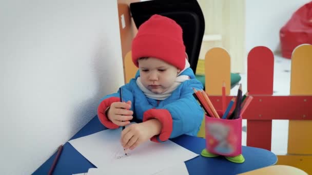 Un niño pequeño calurosamente vestido en la sala de juegos dibuja lápices en el álbum . — Vídeos de Stock