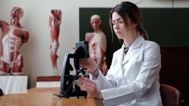 Žena sedí za mikroskopem, na pozadí mannequinů, aby prostudování struktury lidí. — Stock video