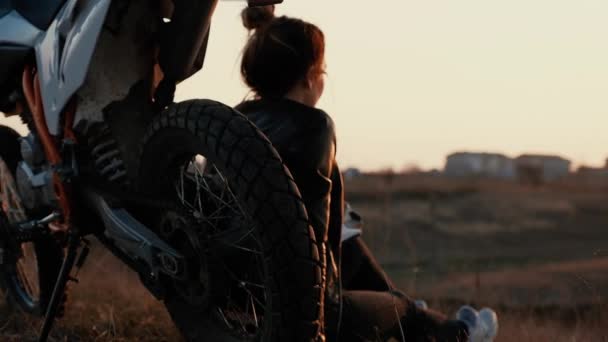 Una joven se sienta al lado de su motocicleta y admira la puesta de sol . — Vídeos de Stock