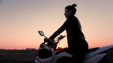 Genç bir kız bir motosiklet astride oturur ve güzel gün batımını izler.