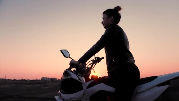 En ung flicka sitter grensle en motorcykel och klockor den vackra solnedgången. — Stockvideo