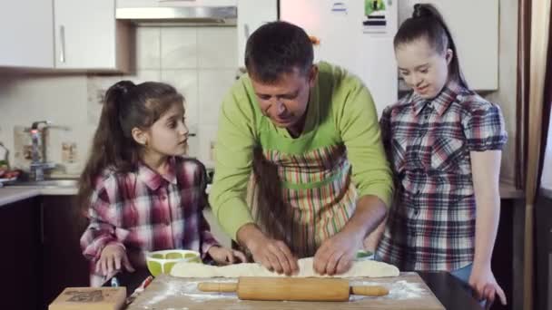 Der Vater zeigt seinen beiden Töchtern, wie man den Teig ausrollt. Eine der Töchter hat Down-Syndrom. — Stockvideo