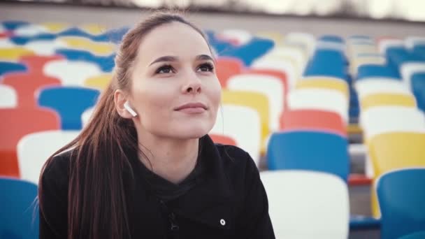 Una ragazza seduta nella fila dello spettatore di un'arena sportiva guarda l'allenamento e ascolta la musica attraverso le cuffie . — Video Stock