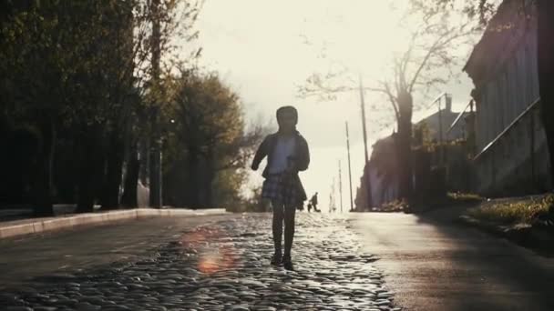 Una niña de nueve años con coletas, corre hacia el pripryshku en la carretera hecha de adoquines en los rayos del sol poniente . — Vídeo de stock