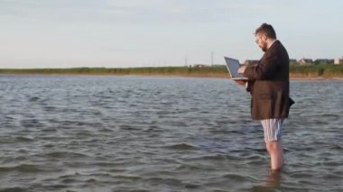 Bir iş kıyafeti ve külot bir adam elinde bir dizüstü bilgisayar ile ayakta ve diz-derin suda.