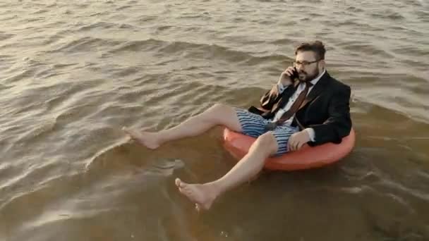 Zakenman zonder broek zittend op een opblaasbare cirkel in het midden van het reservoir en praten op een mobiele telefoon. — Stockvideo