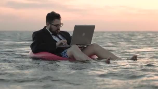 Geschäftsmann arbeitet begeistert am Laptop, wiegt sich auf den Wellen eines Stausees und sitzt auf einem aufblasbaren Kinderzirkel. — Stockvideo