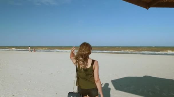 Ένα κορίτσι στέκεται σε μια έρημο παραλία δίπλα από την ομπρέλα παραλίας που γίνεται από καλάμια. — Αρχείο Βίντεο