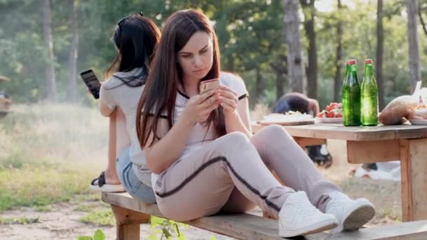 Dvě mladé dívky na pikniku se na svých telefonech dívali s tupým pohledem, zatímco jejich přítel udělal oheň a vaří pro ně. — Stock video