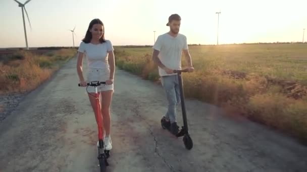 Ein Kerl und ein junges Mädchen bei Sonnenuntergang auf Elektrorollern vor dem Hintergrund von Windkraftanlagen. — Stockvideo