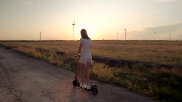 Menina com um cara no pôr do sol entre parques eólicos passeio em scooters elétricos . — Vídeo de Stock