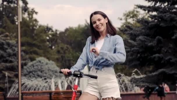 An attractive girl stands next to an electric scooter on the background of a city fountain. — Stock Video