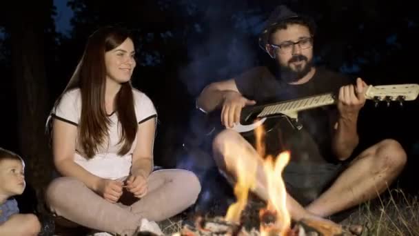 Una divertida compañía con un niño por la noche en el bosque junto al fuego canta canciones a la guitarra . — Vídeos de Stock