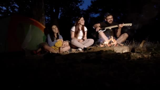 Grupa przyjaciół z dzieckiem śpiewają piosenki na gitarze, relaksując się ogniem. — Wideo stockowe