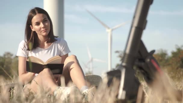 Dziewczyna entuzjastycznie czyta książkę na otwartym powietrzu obok jej skuter elektryczny na tle farm wiatrowych. — Wideo stockowe