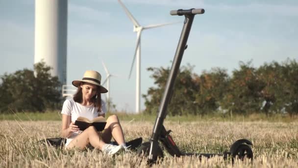 Una chica con un sombrero elegante en la estepa está leyendo un libro, un scooter eléctrico está de pie cerca, en el fondo es un parque eólico . — Vídeos de Stock