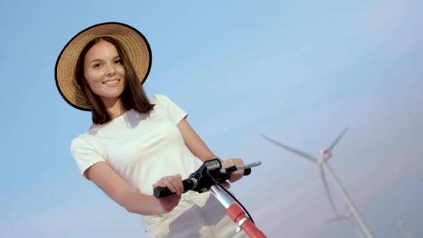 Retrato de una niña en un elegante sombrero de paja con un scooter eléctrico sobre un fondo de parques eólicos . — Vídeos de Stock