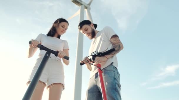 Ein Kerl zeigt seiner Freundin vor dem Hintergrund eines nahe gelegenen Windgenerators die Steuerung eines Elektrorollers. — Stockvideo