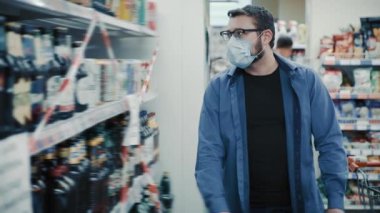 Maskeli adam dükkanda kırmızı ve beyaz kurdeleli biraya bakıyor.