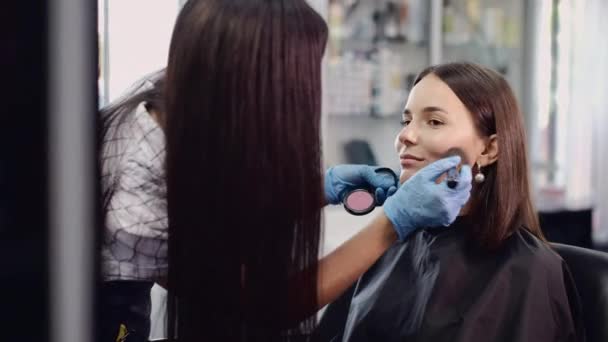 Artista em máscara protetora aplica maquiagem no rosto do cliente da menina — Vídeo de Stock