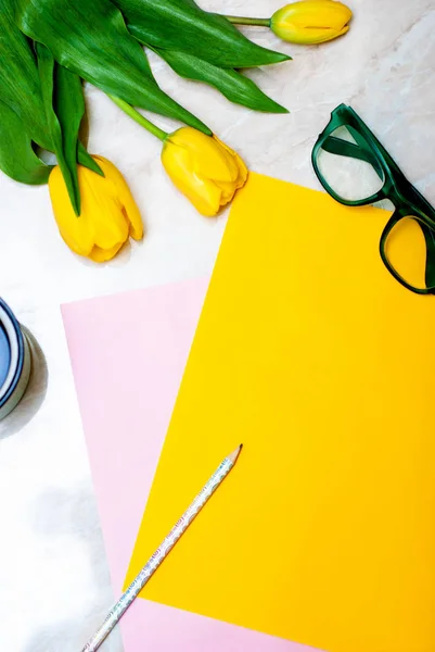 Yellow tulip, coffee, green glasses and colorful paper on marble background