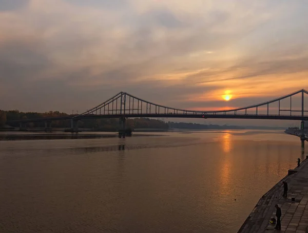 日出时在第聂伯罗彼得罗夫斯克河上的行人桥 太阳在水中反射 人们正在河堤上钓鱼 秋天的风景 乌克兰基辅 — 图库照片