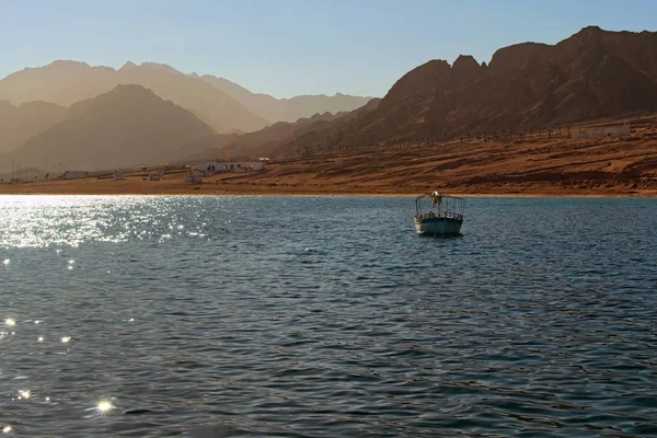 Red Sea in a calm weather. Boat moored near coast into the sea. Beach with some buildings, sand. High mountains at the background. Landscape at sunny hot day. Summer vacation concept. Dahab, Egypt.