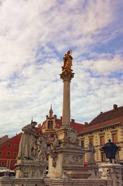 Bulutlu gökyüzüne karşı ortaçağ Veba Sütunu manzarası. Arka planda eski renkli bir bina var. Slovenya 'nın Maribor şehrindeki Rotovz Belediye Meydanı
