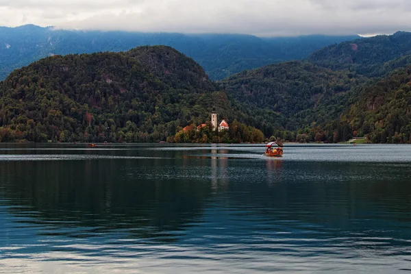 Scenic esti kilátás zarándoklat templom Nagyboldogasszony a kis szigeten a felhős napon. A turistákkal teli hajó visszatér a folyópartra. Bled-tó, Felső-Carniolan régió, Szlovénia — Stock Fotó