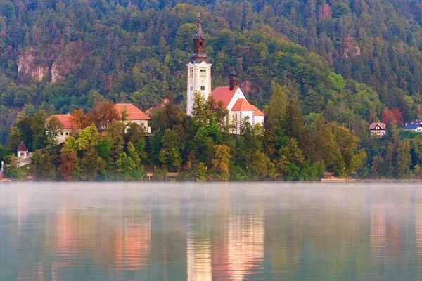 Festői kilátás a Bled-tóra és a Mária mennybemenetele zarándoklati templomra a kis szigeten őszi erdővel szemben. Reggeli köd a víz felszínén. Utazási és turisztikai koncepció. Szlovénia — Stock Fotó