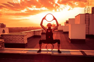Orta yaşlı bir adam yogada tekerlek kullanmakla meşgul. Gün batımında çatıda yoga..